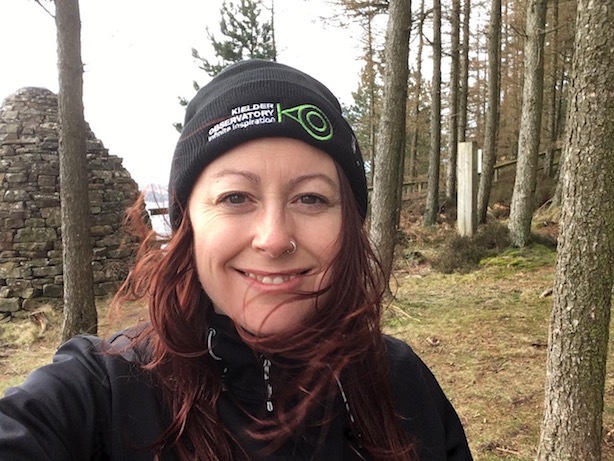 A selfie of me, a white woman with long hair, standing in a forest. I am wearing a black jacket and a beanie hat with a logo for the Kielder Observatory.